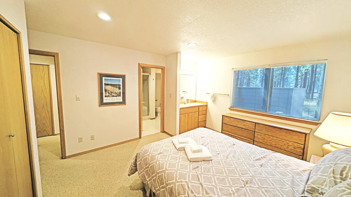 A peaceful bedroom with a bed featuring patterned bedding, a wooden dresser, framed artwork, and a window pouring in natural light with serene outdoor views. The setting feels restful and private.