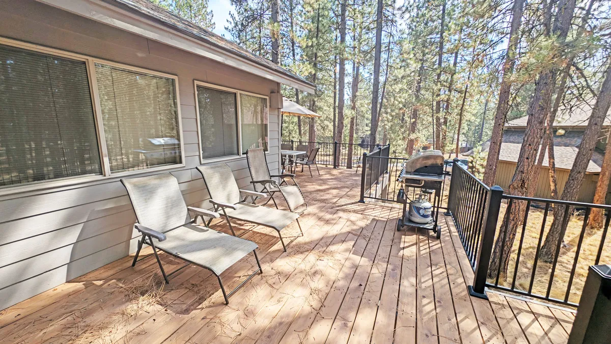 A serene outdoor deck space equipped with comfortable chairs and a grill, set against a backdrop of lush trees. The scene invites guests to enjoy barbecuing, sunbathing, or socializing outdoors in a peaceful setting.