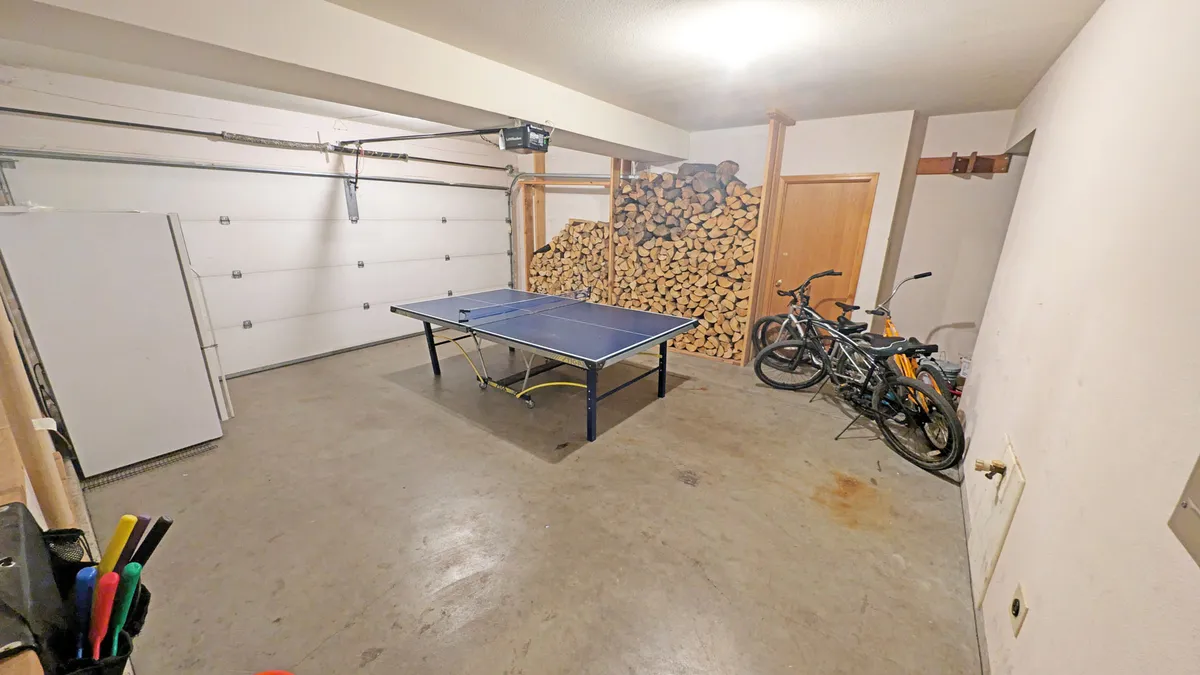 A bright and relaxed garage or game area featuring a ping pong table set up for play, a stack of firewood along one wall, and several bicycles stored neatly by the garage door. The space feels welcoming and ripe for friendly competitions or spontaneous fun.