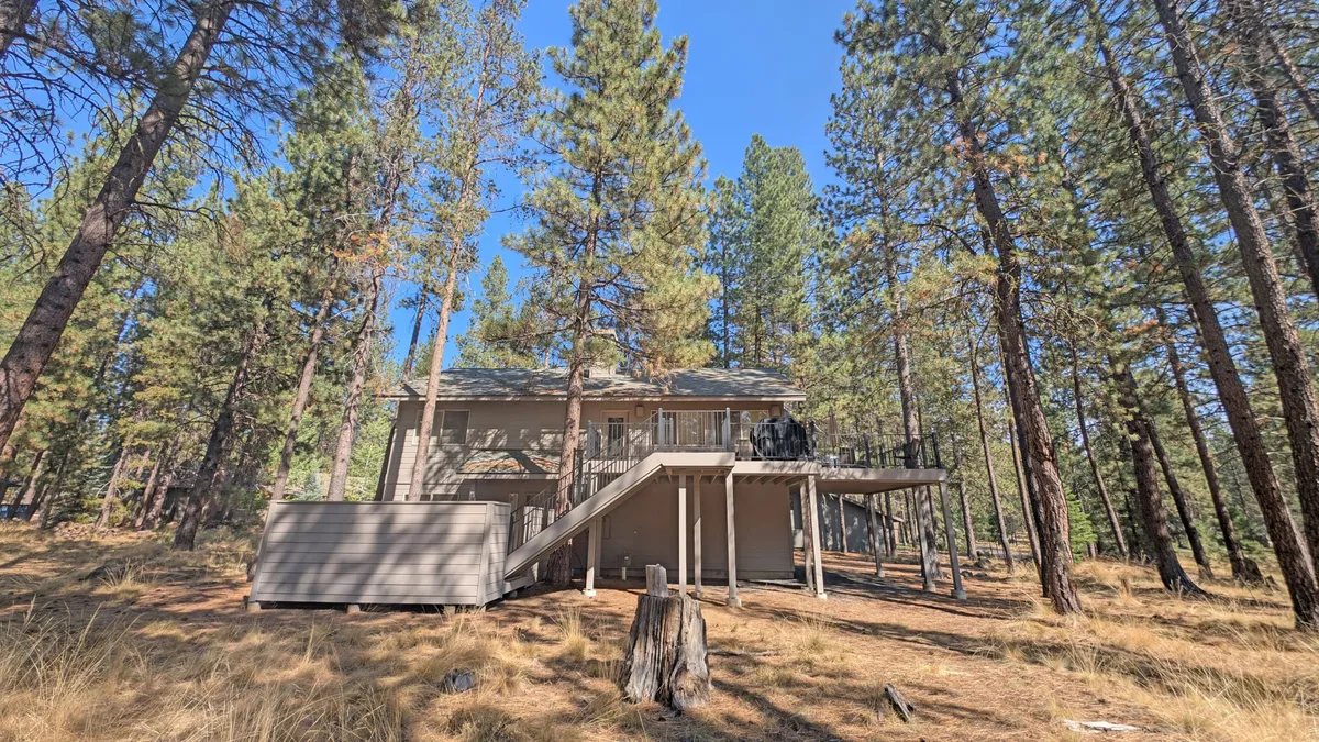 This exterior image shows the house nestled among tall pine trees, with a spacious deck featuring chairs that invite relaxation. The setting highlights both the home and its natural surroundings, emphasizing outdoor living and the ease of access to nearby trails and parks.