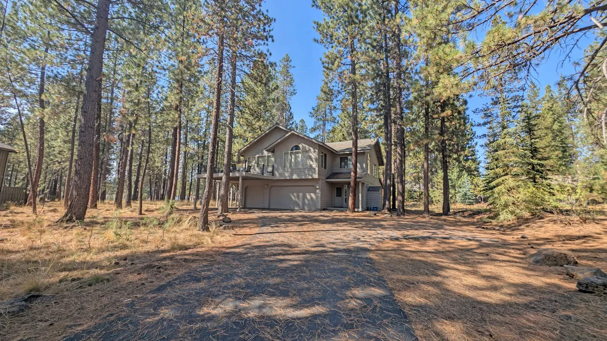 A wide-angle view of the house framed by towering pine trees, with a welcoming driveway leading up and the balcony visible. The scene suggests opportunities for gathering, outdoor activities, and appreciating the surrounding forest.