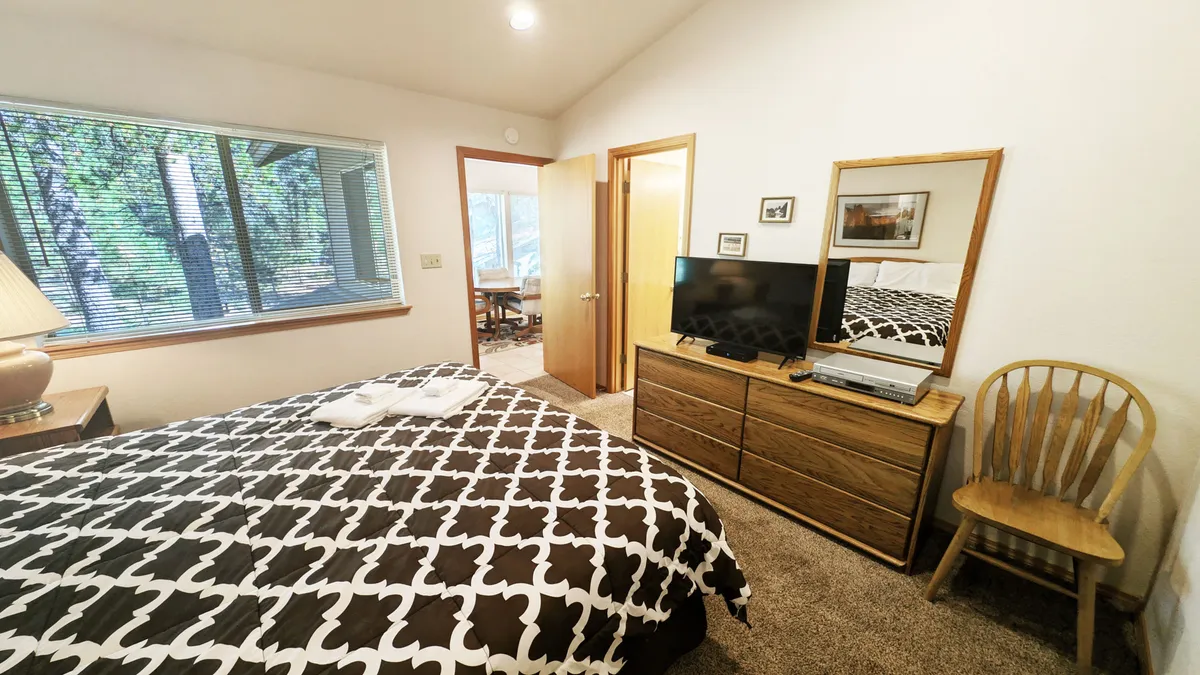 A bright and comfortable bedroom featuring a bed with patterned bedding, a large window that lets in ample natural light, a wooden dresser with a flatscreen TV, and a decorative mirror. The room feels inviting and relaxing, perfect for unwinding.