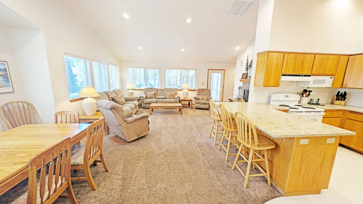 This image shows a bright and inviting interior with large windows, comfortable sofas, a dining table, and a central kitchen island. Natural light pours in, highlighting the seamless connection between the living, dining, and kitchen spaces. The room is set up perfectly for both relaxation and hosting gatherings.
