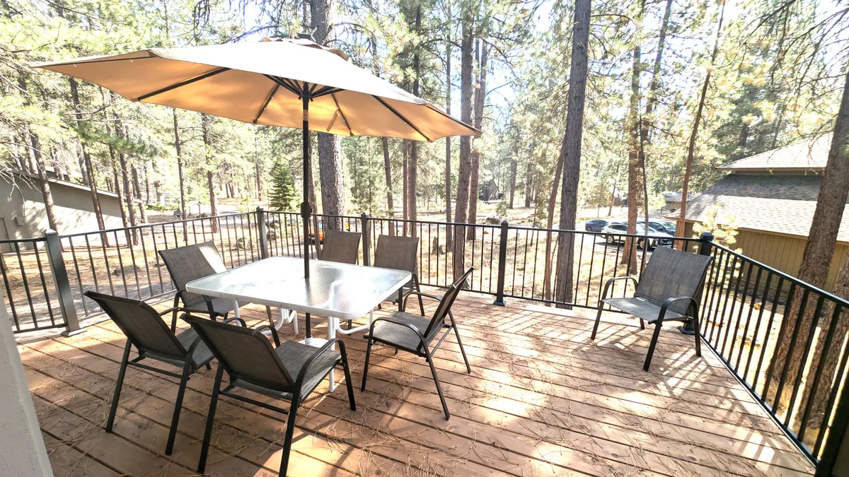 The image captures an outdoor dining setup, including a table and chairs beneath an umbrella, set against a backdrop of trees. The relaxed atmosphere hints at family gatherings, alfresco meals, and the enjoyment of nature just steps from the home.
